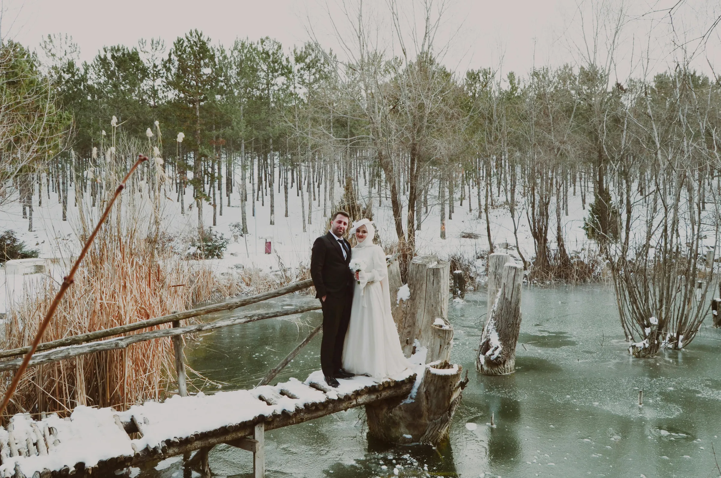 Kar yağışı altında rustik ahşap masa düzenlemesi, altın şamdanlar, bordo güller ve çam kozalakları ile süslenmiş kış düğünü dekorasyonu